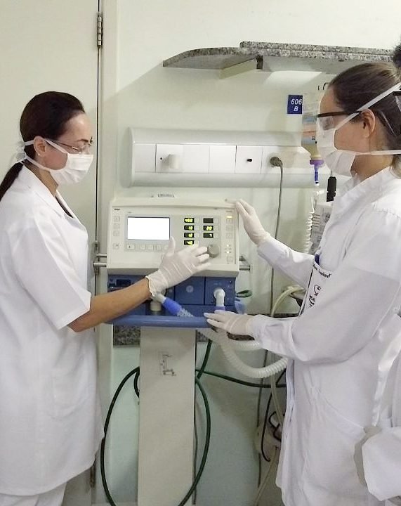 Professora e aluna de Fisioterapia ajustando um ventilador mecânico em ambiente hospitalar